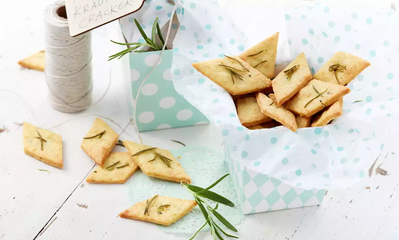 Goldgelbe, rhombenförmige Kräuter-Cracker mit fein strukturierter Oberfläche, ansprechend in gemusterter Schachtel mit Rosmarinzweig arrangiert