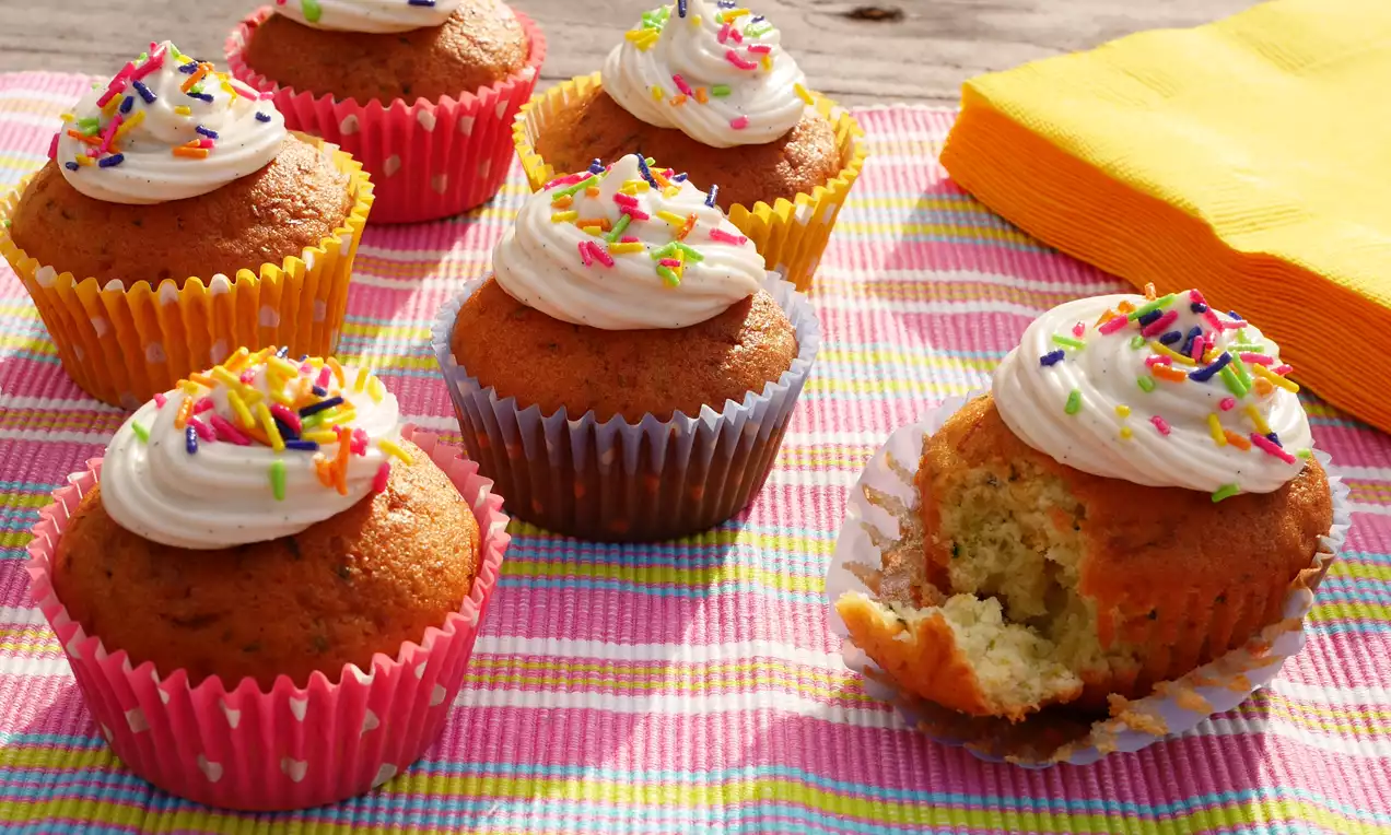 Zitronen-Zucchini-Cupcakes in bunten Papierförmchen mit cremigem weißen Topping und farbigen Streuseln auf gestreifter Tischdecke