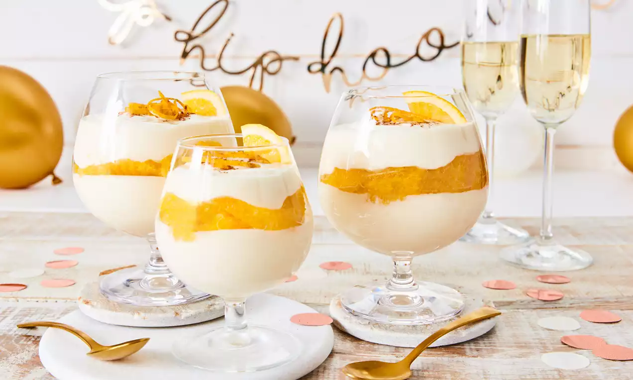 Schichtdessert mit sahnig-cremer Gelb-Weißen layern, Zitruszesten-Topping in bauchigen Gläsern goldene Deko, feierlich.