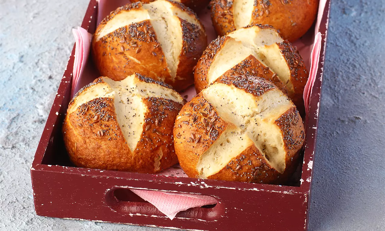 Altın kahverengi ve kabarık laugenbrotchen ekmekleri, üzeri çapraz kesimli ve tohumlarla serpilmiş, kırmızı ahşap tepsi içinde düzenlenmiş