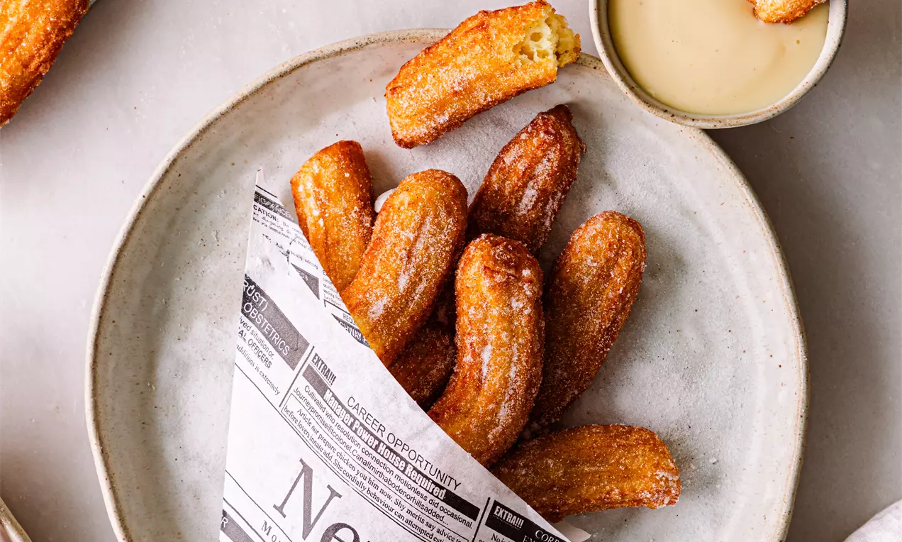 Picture - Golden churros with crisp ridged surfaces wrapped in printed paper cone beside a creamy beige dipping sauce on a pale ceramic plate