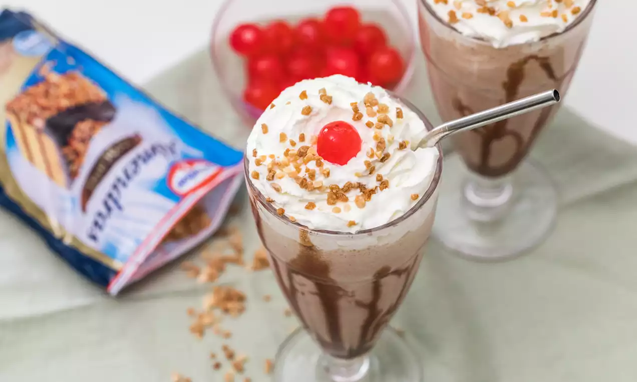 Batido espeso de chocolate marrón decorado con crema blanca, chispas caramelizadas, cereza roja brillante y pajilla plateada en vaso alto
