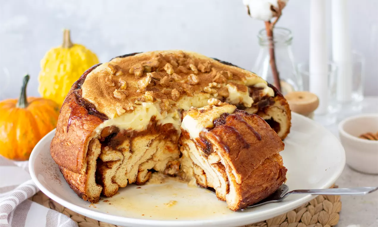 Овален сладкиш с карамелено-кафява коричка, разкошна кремообразна текстура и орехови парченца отгоре, върху бяла чиния с тиква на заден план