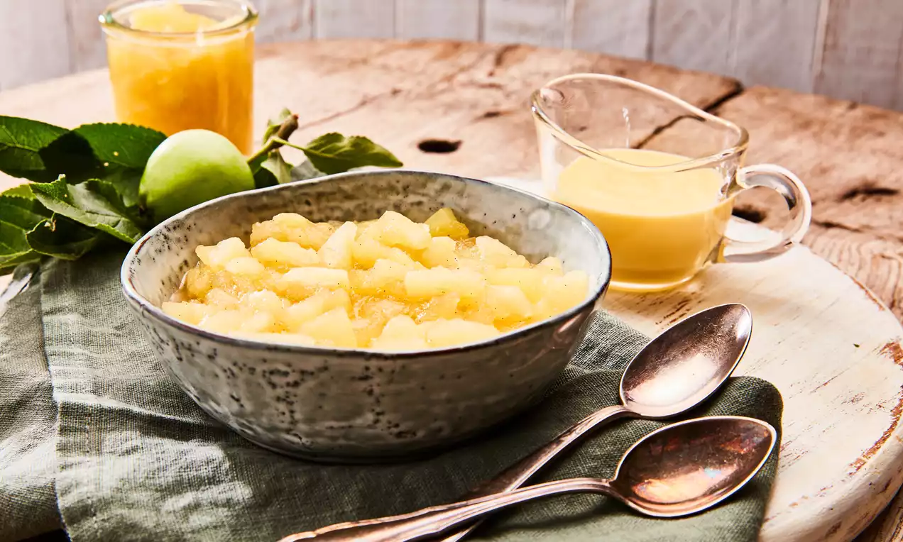 Apfelkompott in einer grauen Keramikschale auf grünem Stoff, daneben zwei Löffel, ein Glas Apfelsauce, Vanillesoße und grüne Blätter