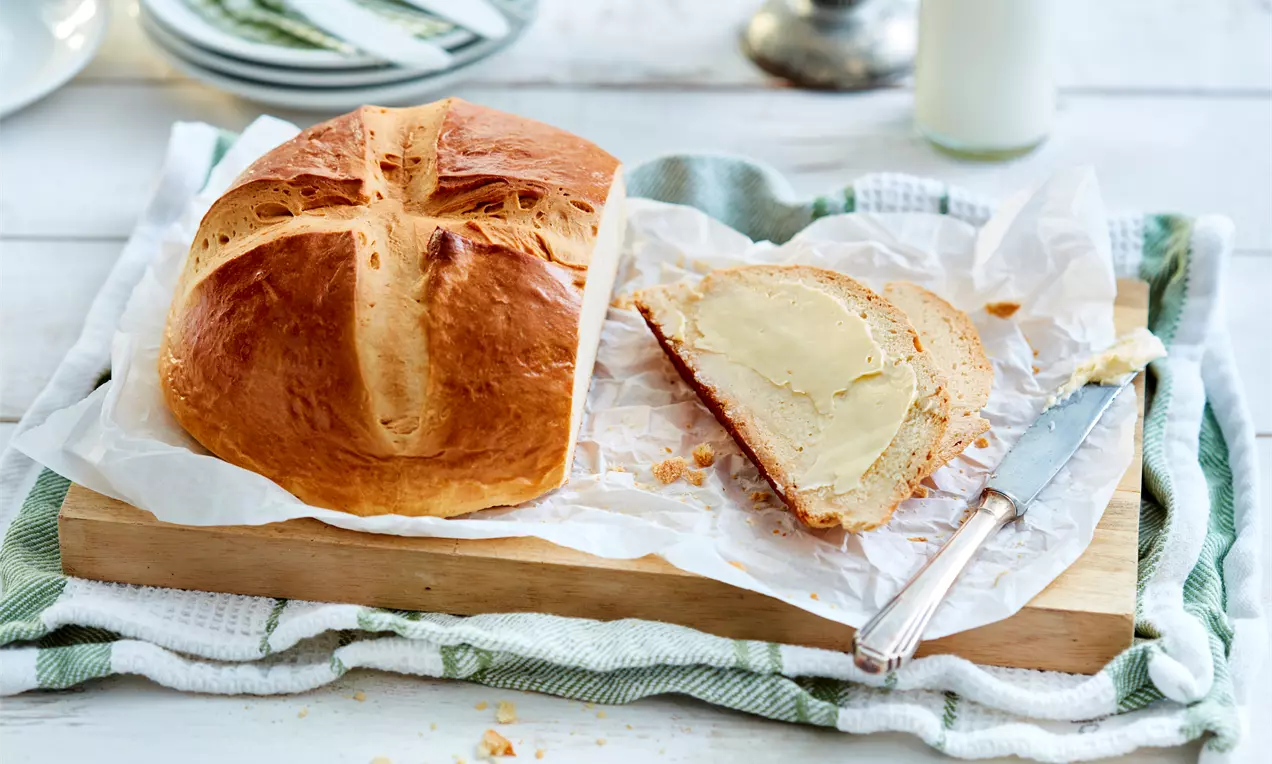 Златисто-кафяво козуначено тесто със загладен ръст и разрези по повърхността, поднесено върху дървена плоскост с нож и резен върху хартия