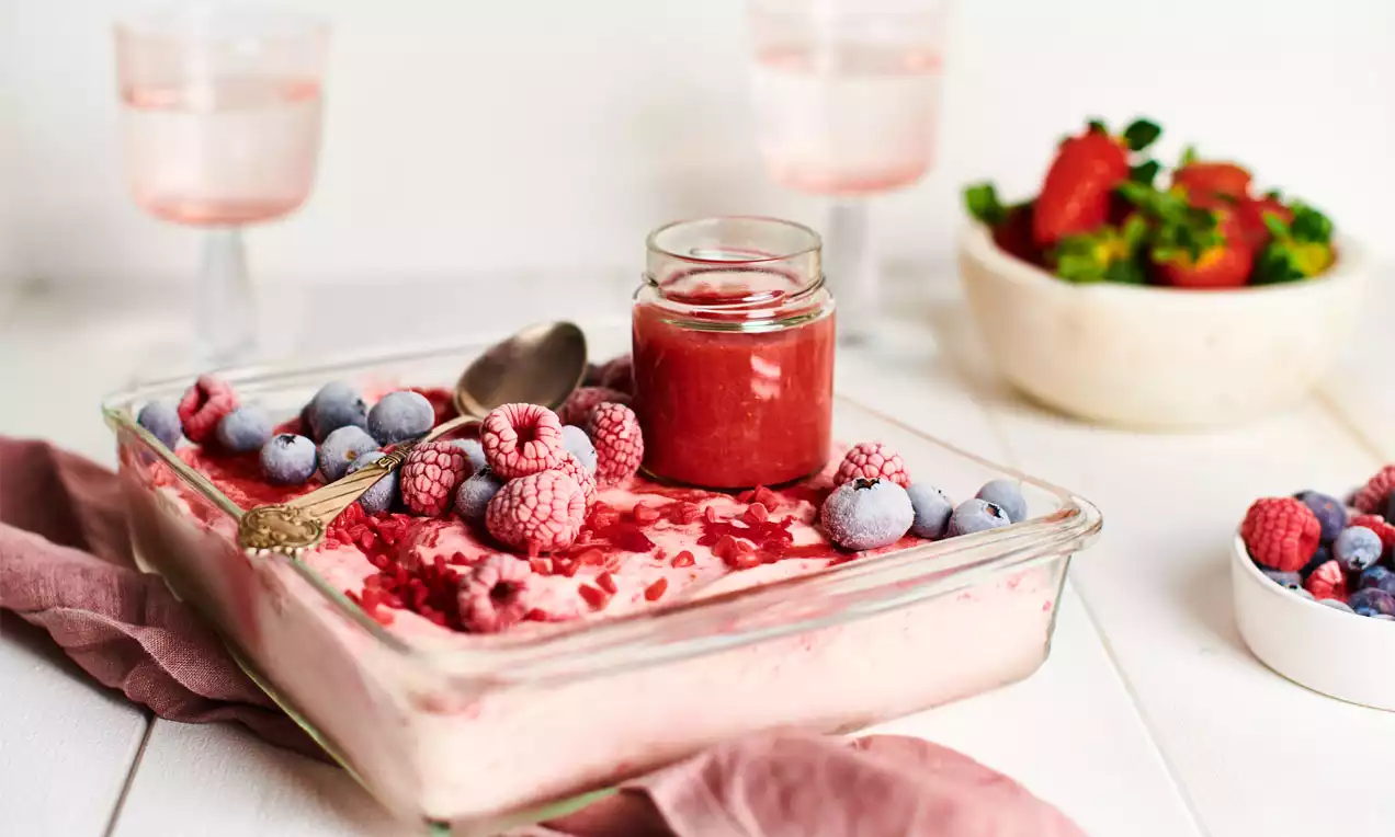 Cremiges rosafarbenes Vanilleeis in einer Glasschale, dekoriert mit gefrorenen Himbeeren, Blaubeeren und einer kleinen Flasche roter Soße