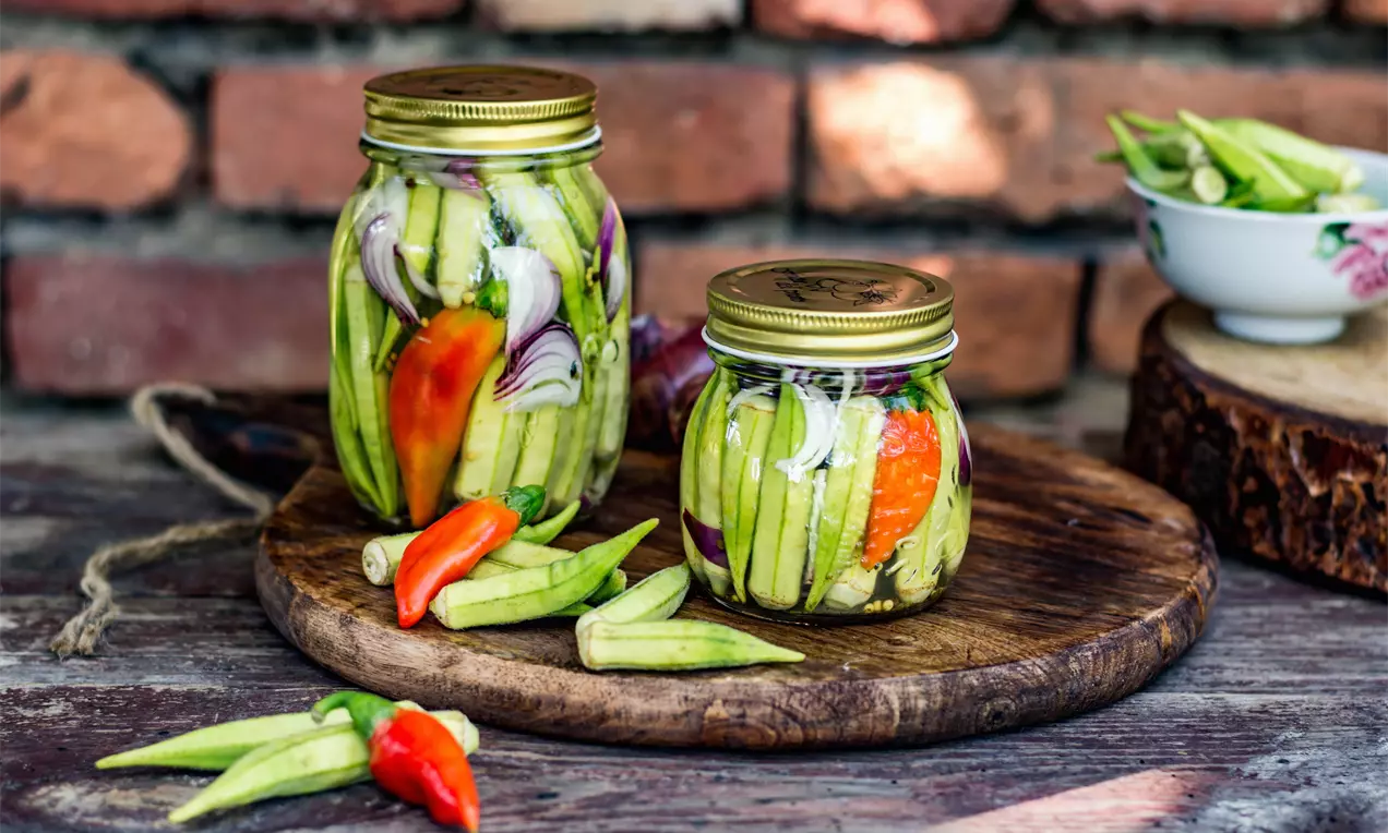 Бамя в буркани с яркозелена текстура, украсена с резени лилав лук и червени чушки, поставена върху дървена плоча пред тухлена стена