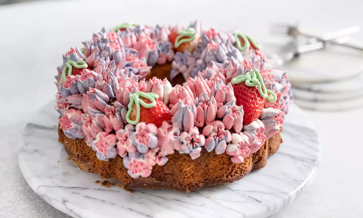 Bundtkaka på marmorfat dekorerad med rosa och lila smörkrämsblommor, röda jordgubbar och gröna blad i kräm, med gafflar i bakgrunden
