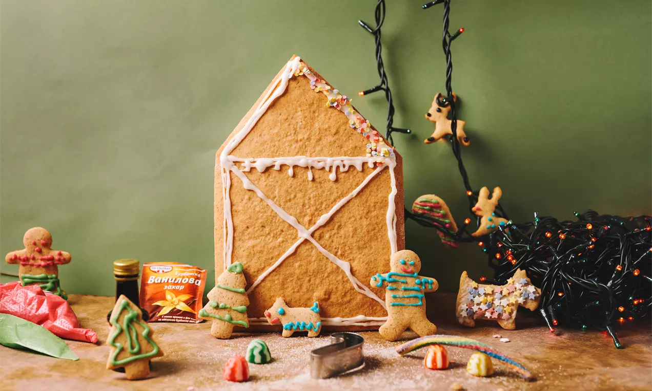 Коледна къщичка от светлокафяво джинджифилово тесто с бели глазурани детайли, заобиколена от цветни бисквити и празнични декорации
