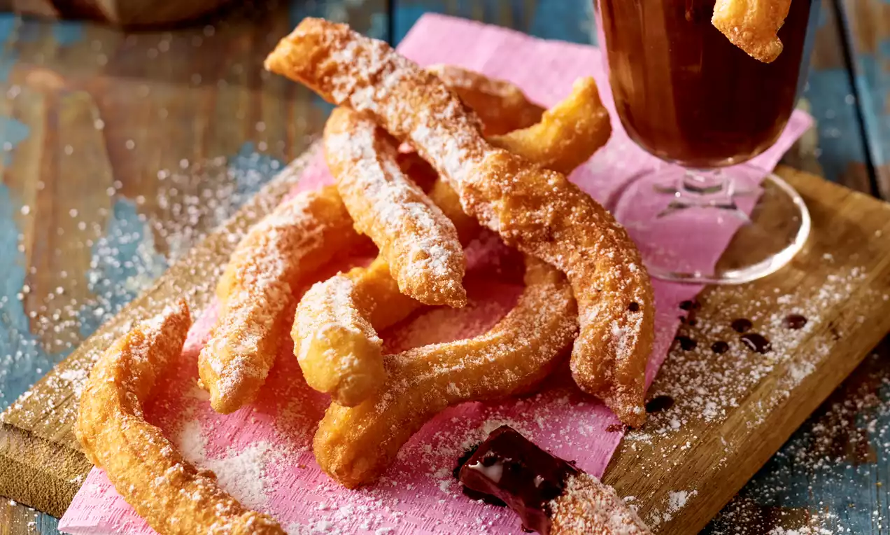 Zlato rjavi ocvrtki podolgovate oblike s hrustljavo površino, posuti s sladkorjem, na rožnatem papirju ob kozarčku goste čokolade