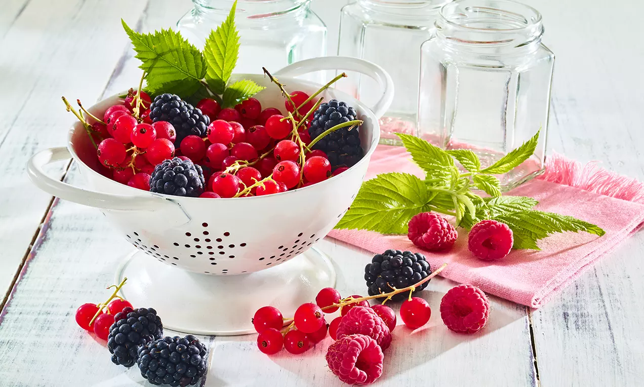 Šťavnaté červené a čierne bobule v bielom kovovom cedidle, obklopené malinami, listami a prázdnymi sklenenými pohármi na drevenom stole