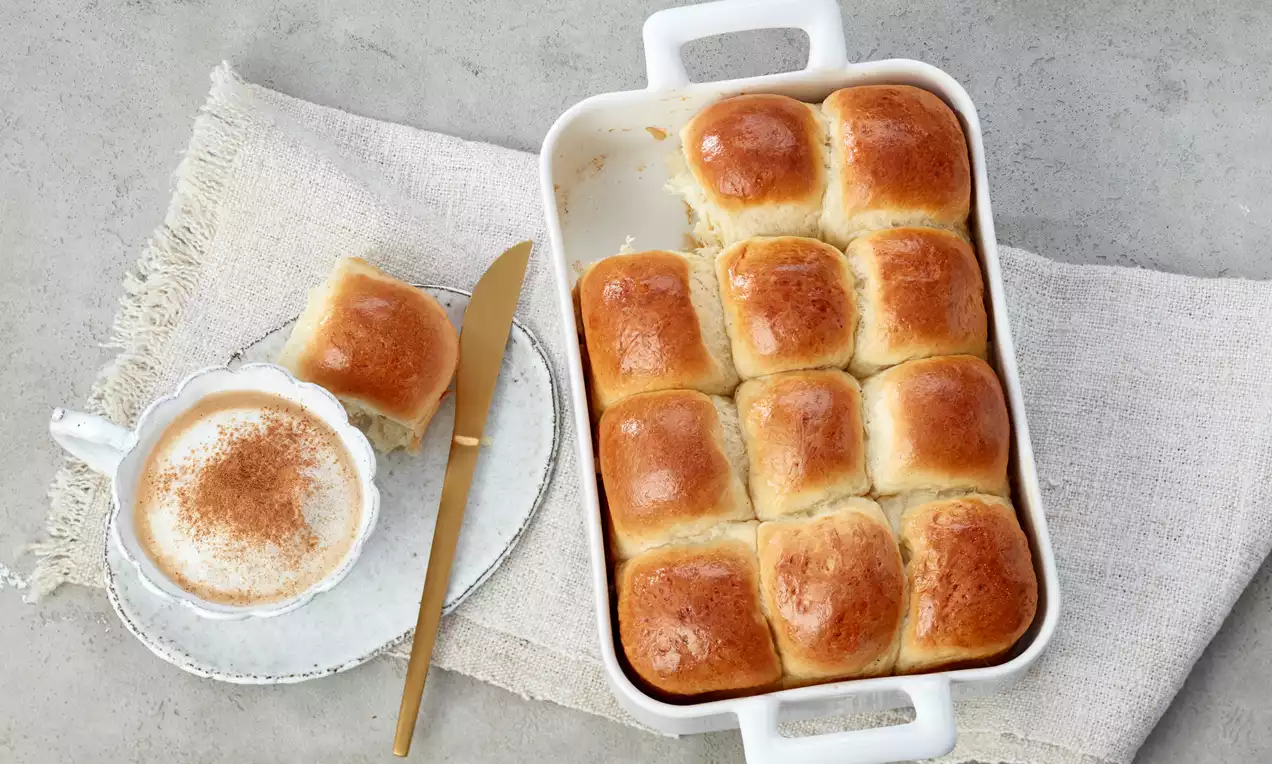Goldbraune, glänzende Milchbrötchen in einer weißen Auflaufform, daneben Tasse mit Milchschaum und Zimt auf angerautem, hellem Stoff
