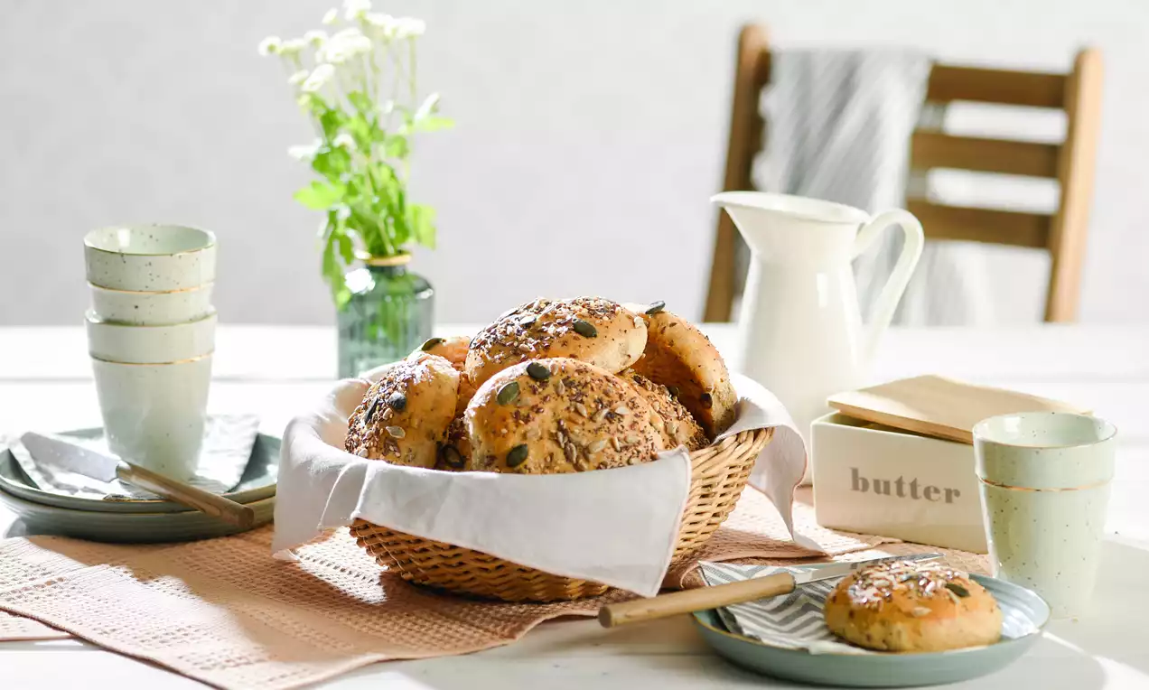 Goldbraune Körnerbrötchen mit knackiger Saatenkruste in einem Korb mit Stofftuch auf hellem Tisch mit Geschirr, Butterdose und Milchkanne
