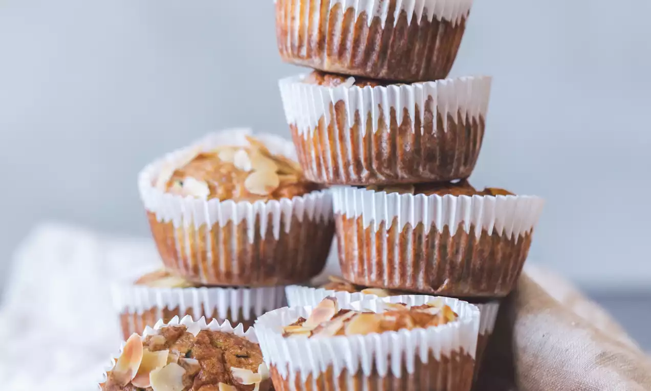 Muffins dorados con superficie coronada por láminas de almendra, envueltos en cápsulas blancas y apilados sobre un fondo azul suave