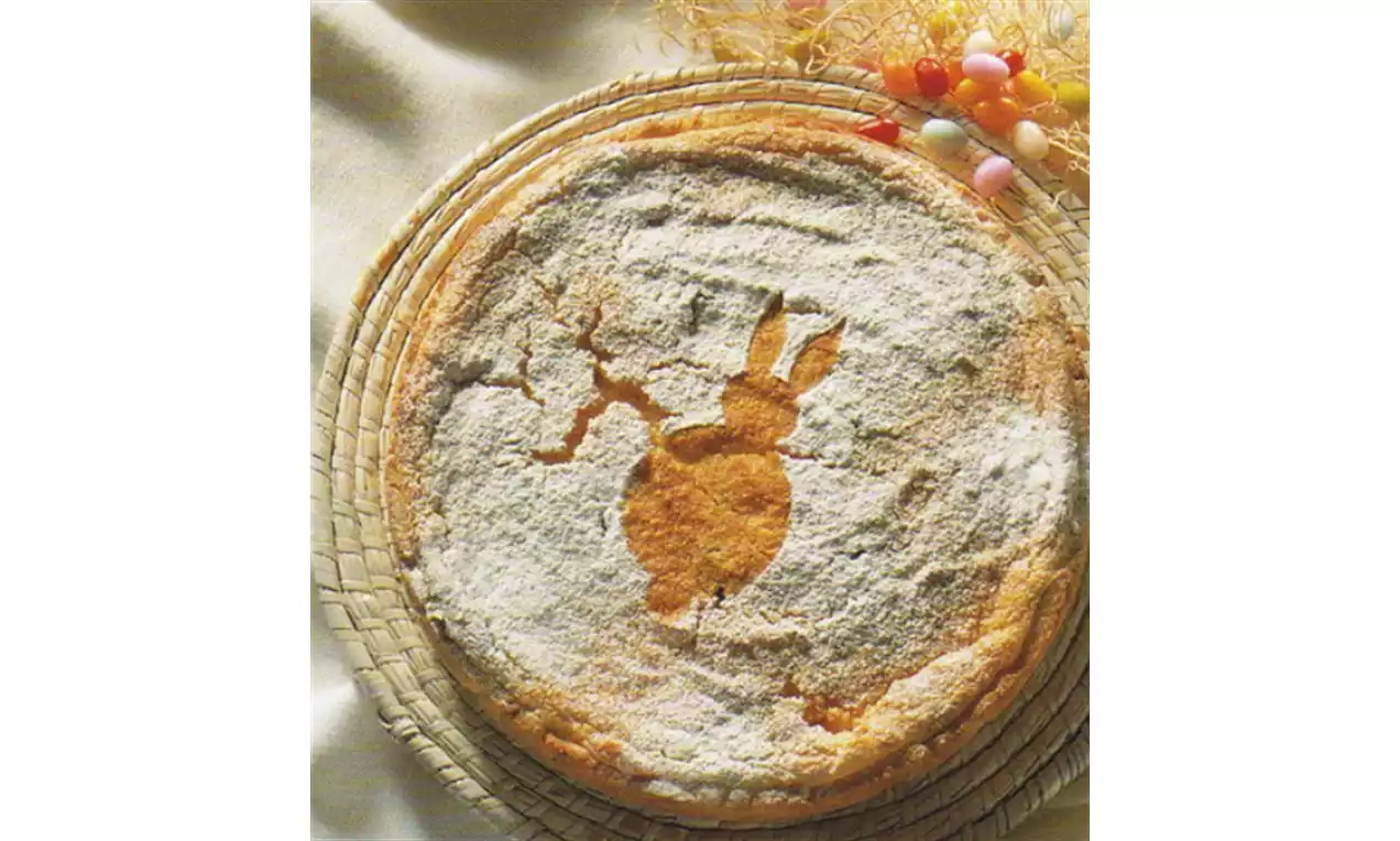 Goudbruine ronde taart met een konijntjesvorm in poedersuiker op een rieten ondergrond, omgeven door pastelkleurige paaseitjes
