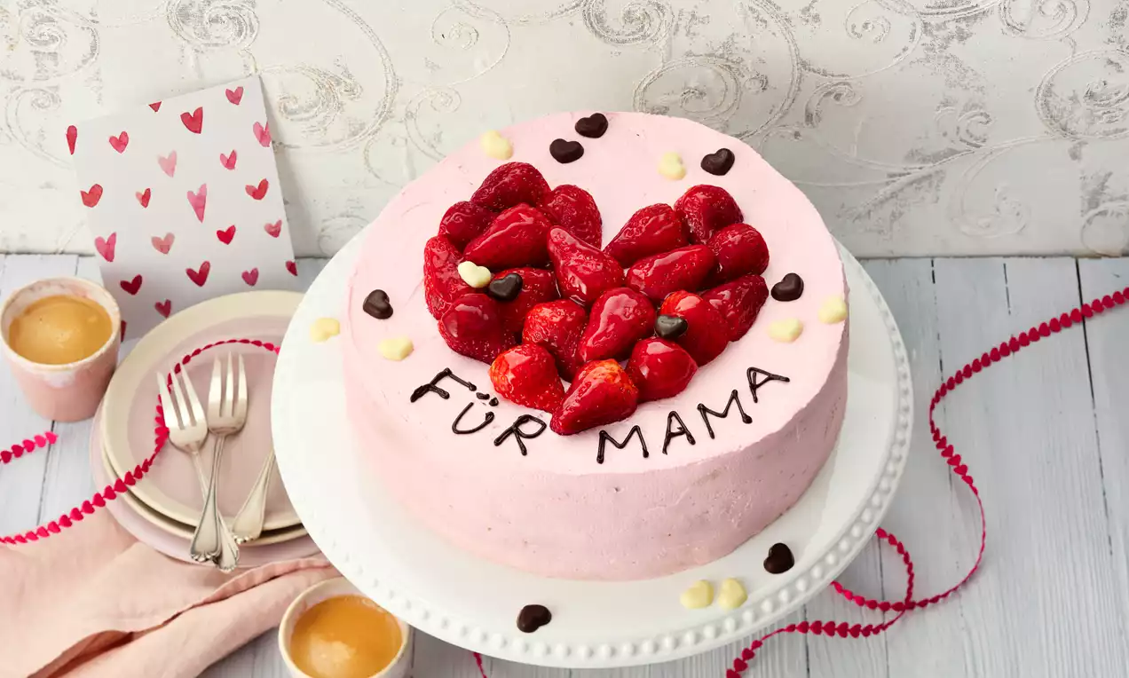Rosafarbene Erdbeertorte mit Fruchtherz aus glasierten Erdbeeren und Schokoladendekor auf weißer Tortenplatte, umgeben von Muttertagsdetails