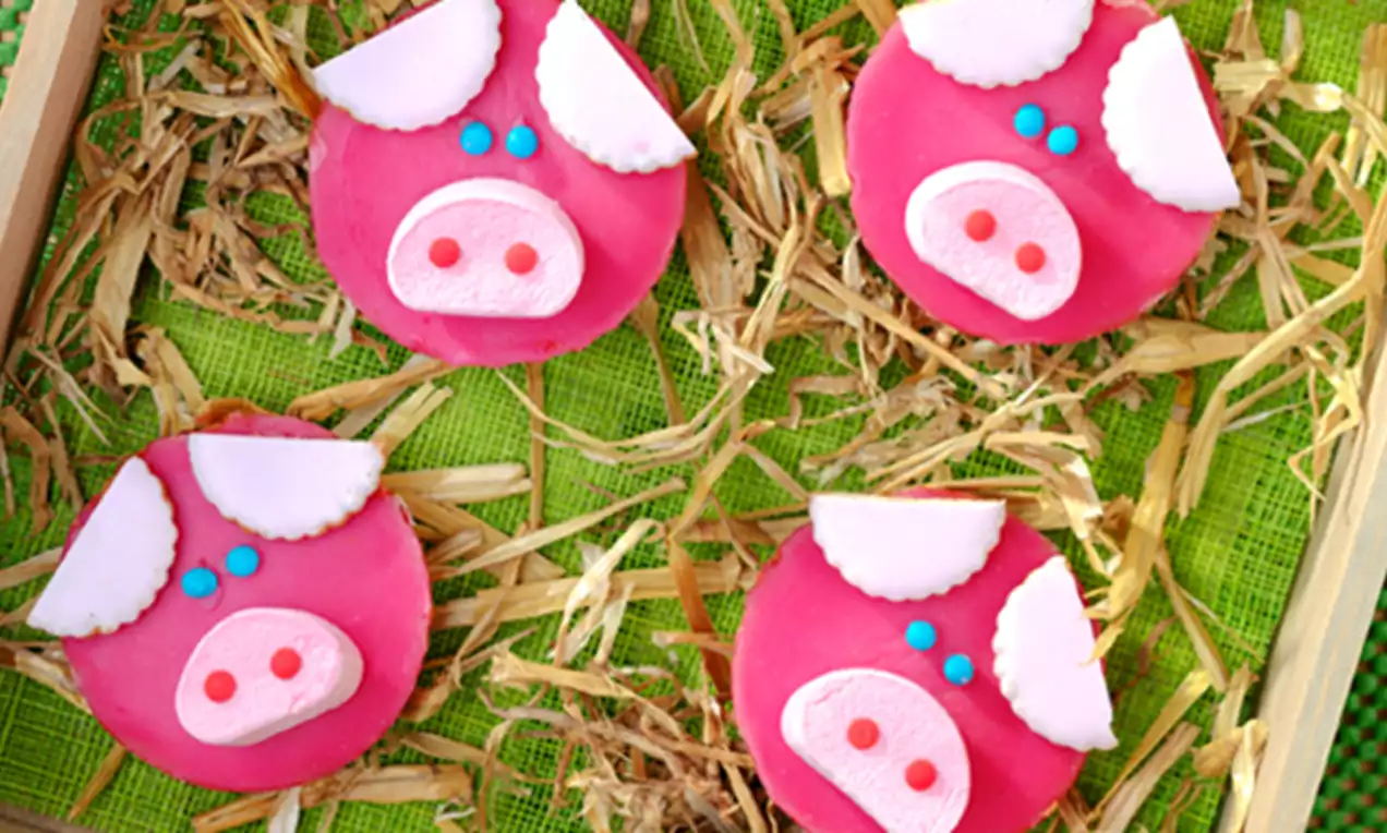 Galletas decoradas con caras de cerditos rosa brillante, detalles blancos y azules, sobre un lecho de paja y fondo verde vibrante