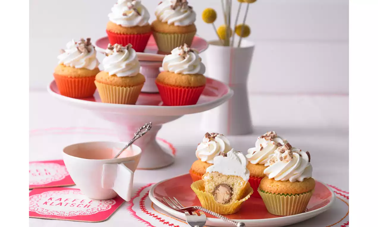 Mini-Muffins mit heller Füllung und lockerer Cremehaube, teils auf einer rosa Tortenplatte, teils auf einem Teller mit Gabel arrangiert