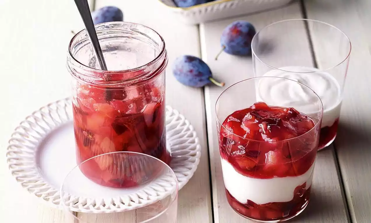 Rotviolettes Birnen-Zwetschgen-Kompott mit Zimt in einem Glas auf weißem Teller neben durchsichtigen Dessertgläsern mit Sahne und Früchten