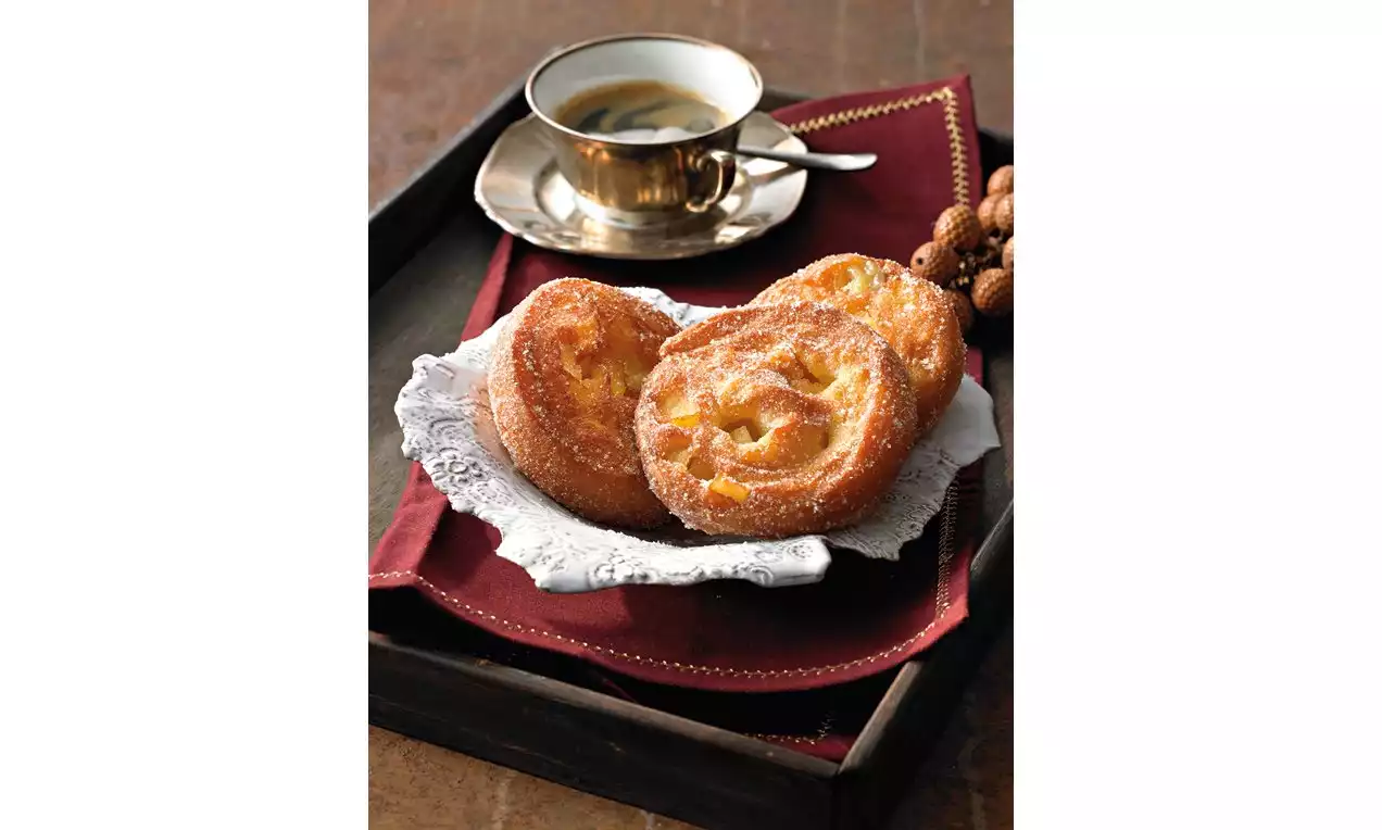 Omenamunkit pehmeän kullankeltaisina, sokerilla kuorrutettuina, pitsireunaisella lautasella punaisella liinalla kahvikupin rinnalla