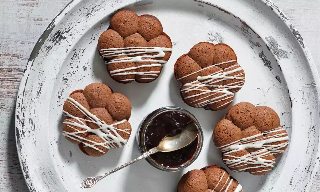 Tmavě hnědé kakaové sušenky ve tvaru květů s jemnými rýhami, zdobené bílou cukrovou polevou, uspořádané na oprýskaném bílém talíři
