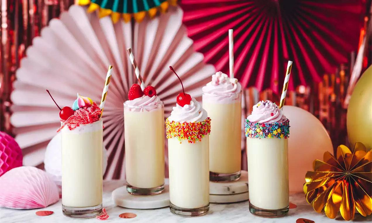 Vanilkové nápoje v vysokých sklenicích s dekorativní šlehačkou, barevným posypem a třešněmi na mramorovém povrchu, obklopené papírovými dekoracemi