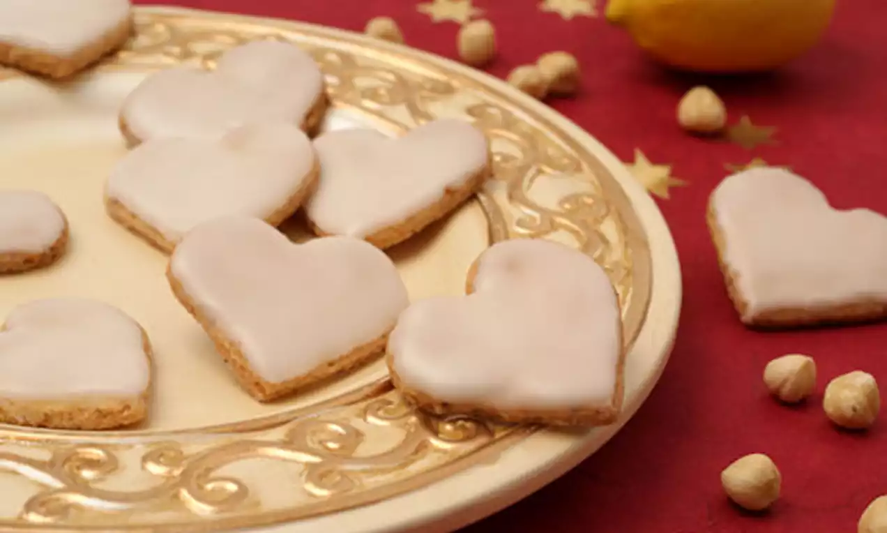 Herzförmige Haselnussplätzchen mit glatter Zitronenglasur auf verziertem goldenen Teller, umgeben von Haselnüssen und einer Zitrone auf rotem Tischtuch