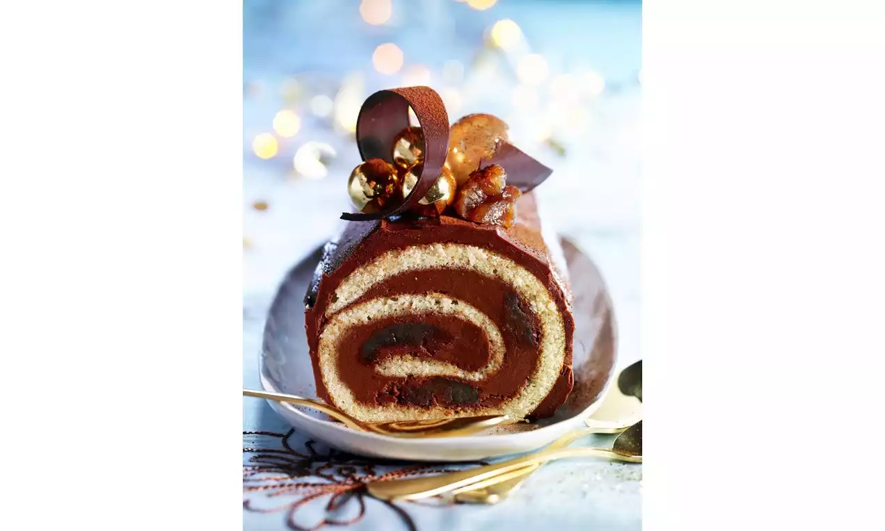 Chocoladebûche met glanzende bruine topping, spiraalvormige vulling, chocoladekrullen en noten op een wit bord met gouden bestek