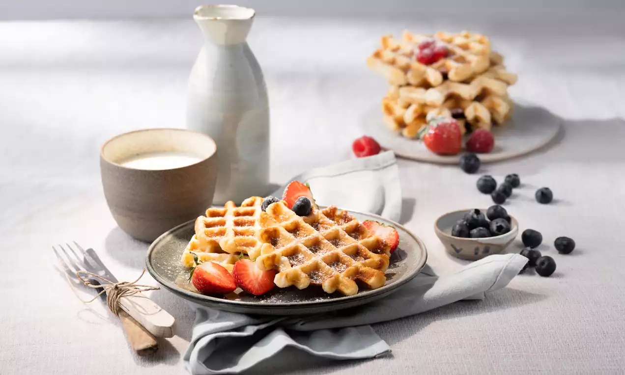 Goudbruine wafels met poedersuiker, omringd door blauwe bessen en aardbeien op een donker bord met melk in een karaf en kop ernaast