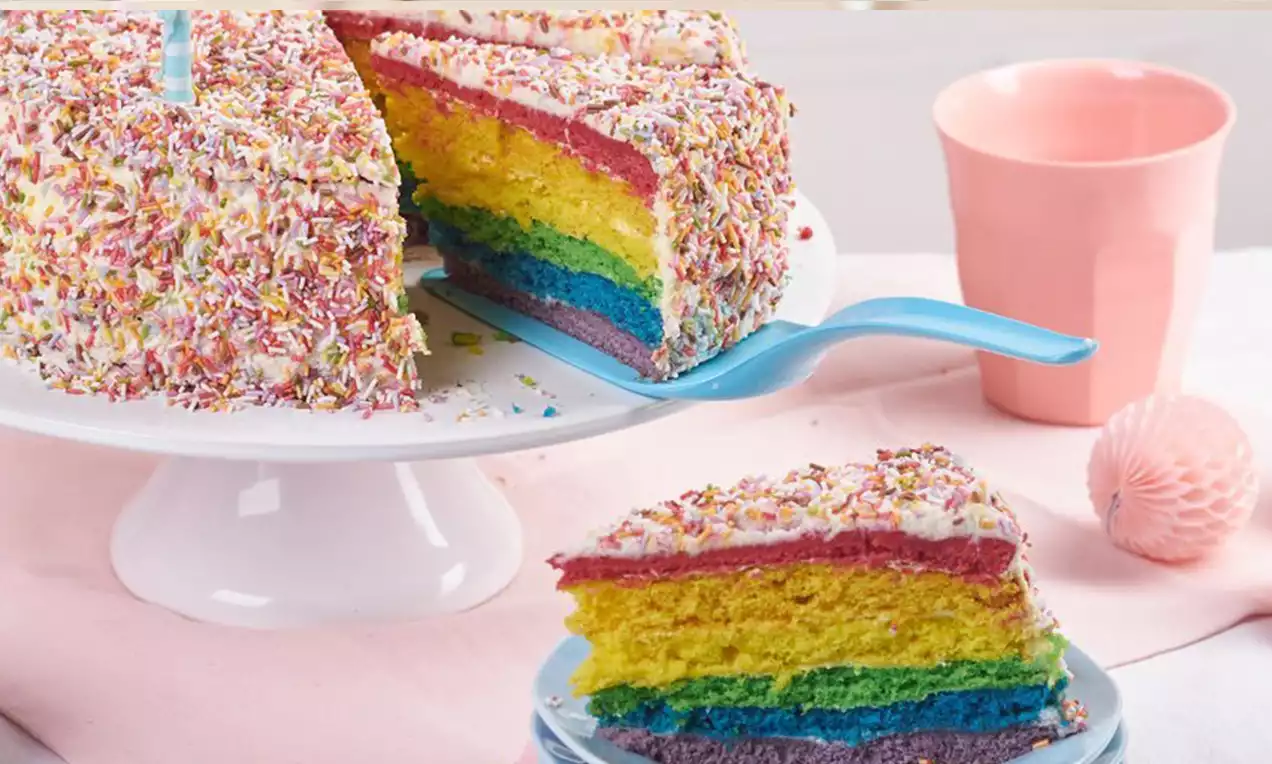 Torta arcobaleno a strati dai colori vivaci con glassa bianca e granella colorata su alzata bianca, servita con fetta su piattino azzurro