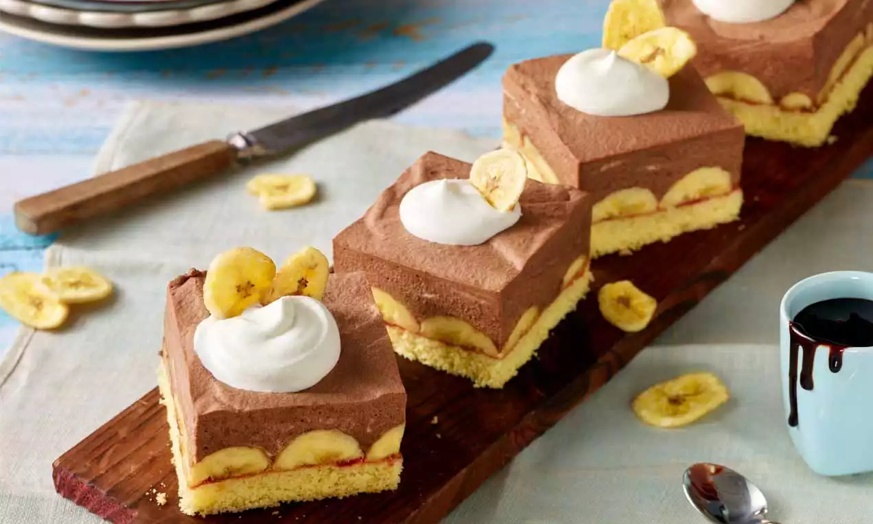 Kocke od mekane žute kore s kriškama banane, čokoladnim mousseom, bijelom hrpicom šlaga i komadićima sušene banane na drvenoj podlozi