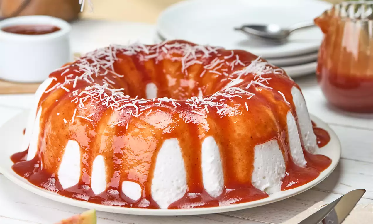 Pudim em forma de anel com textura aerada branca coberto por calda avermelhada brilhante e fios finos de coco em um prato branco