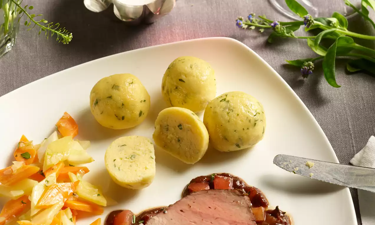Grießknödel, rund und hellgelb mit grünen Kräuterpunkten, weich texturiert, neben Gemüse und Braten auf einem weißen Teller angerichtet