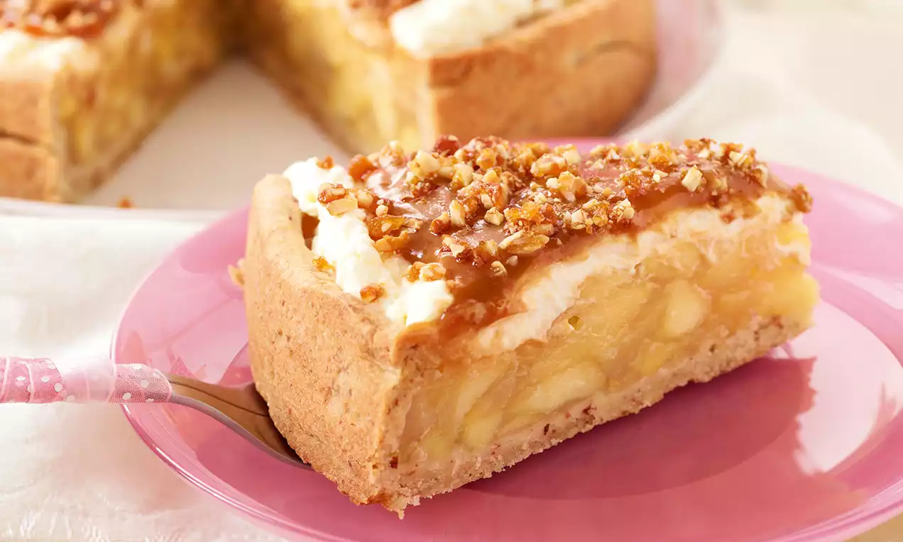 Ein Stück Apfelkuchen mit karamellbrauner Sauce, gehackten Nüssen und heller Sahne auf rosa Teller mit pastellfarbener Gabel drapiert