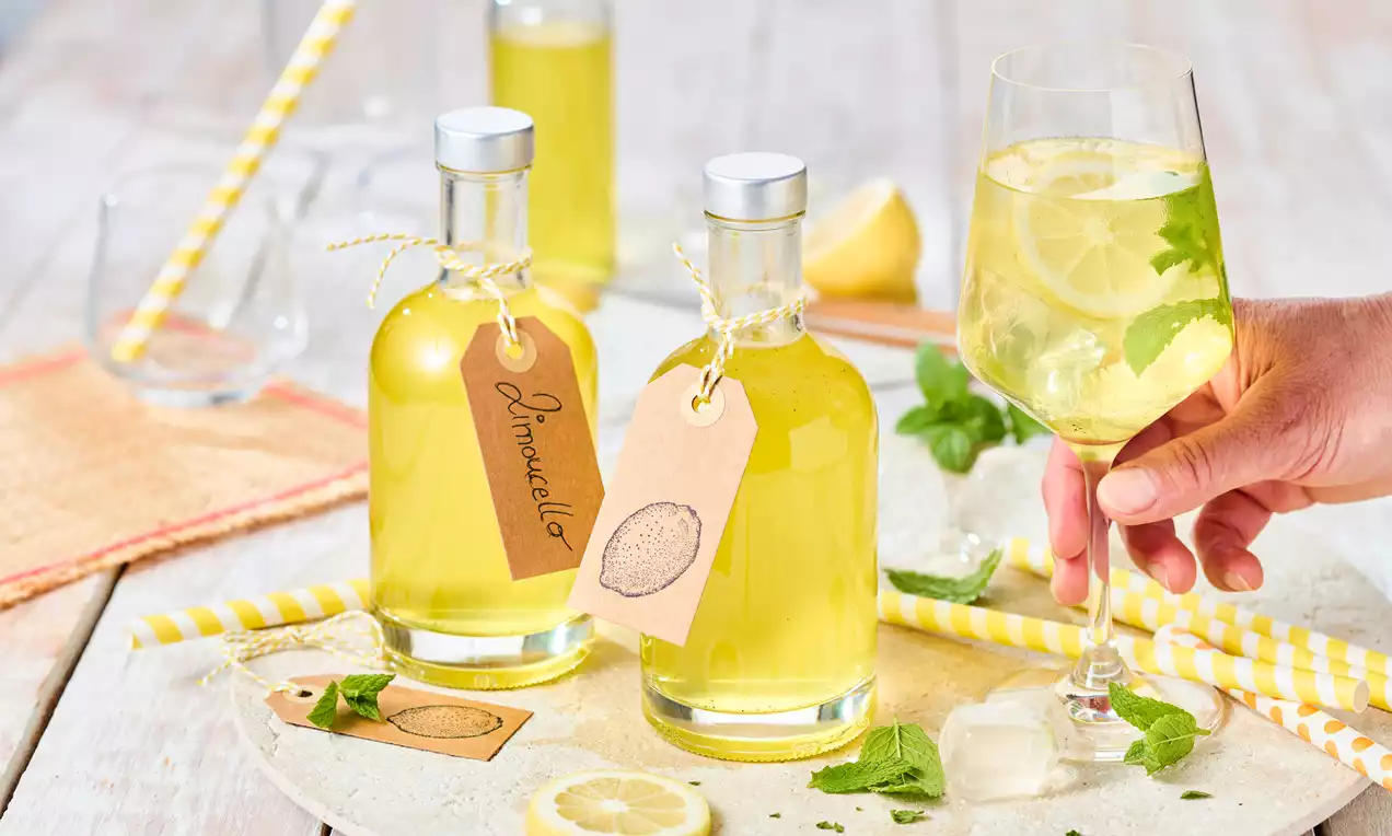 Flaschen mit gelbem Limoncello und handgeschriebenen Etiketten neben einem Glas mit Zitronenscheiben, Minze und Eiswürfeln auf hellem Tisch