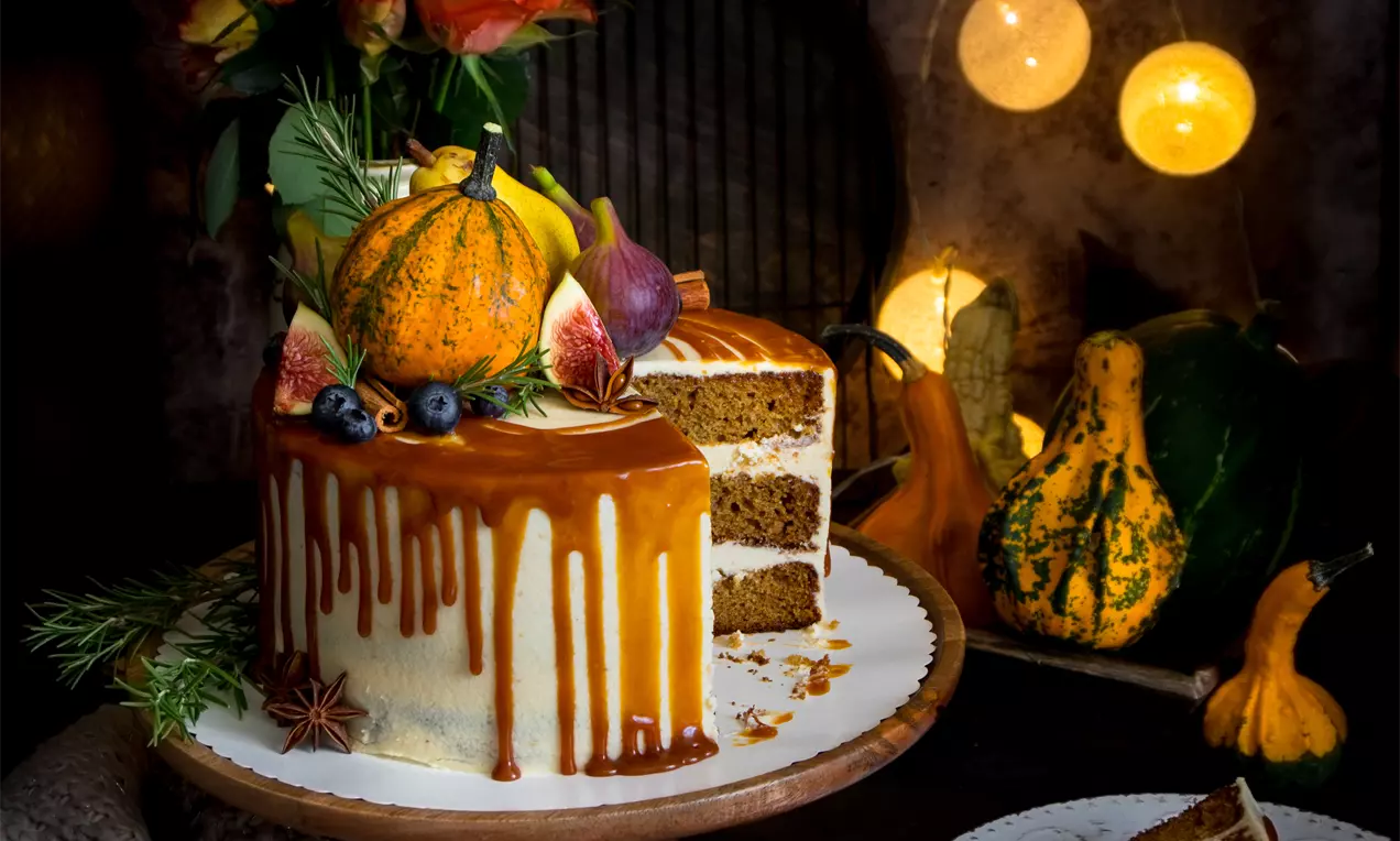 Тиквена торта с топъл оранжев карамелен глазур, слоеве светъл блат и крем, декорирана с мини тиква, смокини, боровинки и клонки върху дървена поставка