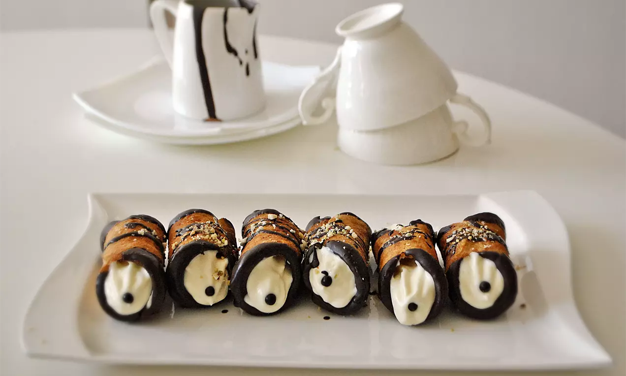 Шест каноли с шоколадово покритие, крем от вътрешността и поръска, подредени в редица върху бяла чиния с фон от чаши и каничка