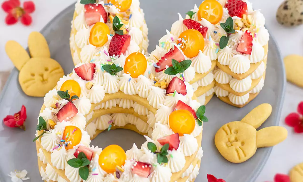 Torta u obliku zeca od prhkog tijesta slojevitih ušiju i glave, ukrašena šlagom, jagodama, mini cvjetovima i kriškama kumkvata na sivom tanjiru