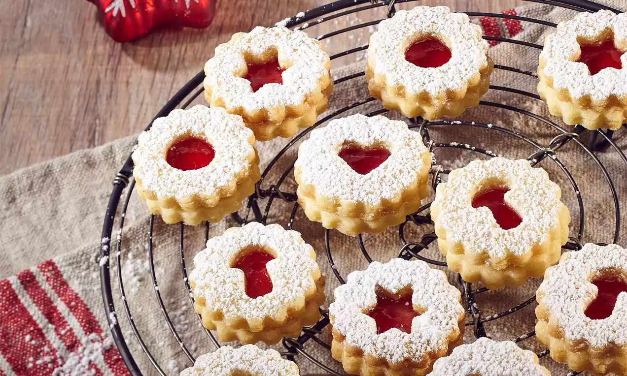 Kulaté jemně zlatavé sušenky s vykrojeným středem plněným červenou marmeládou, posypané moučkovým cukrem na černé mřížce