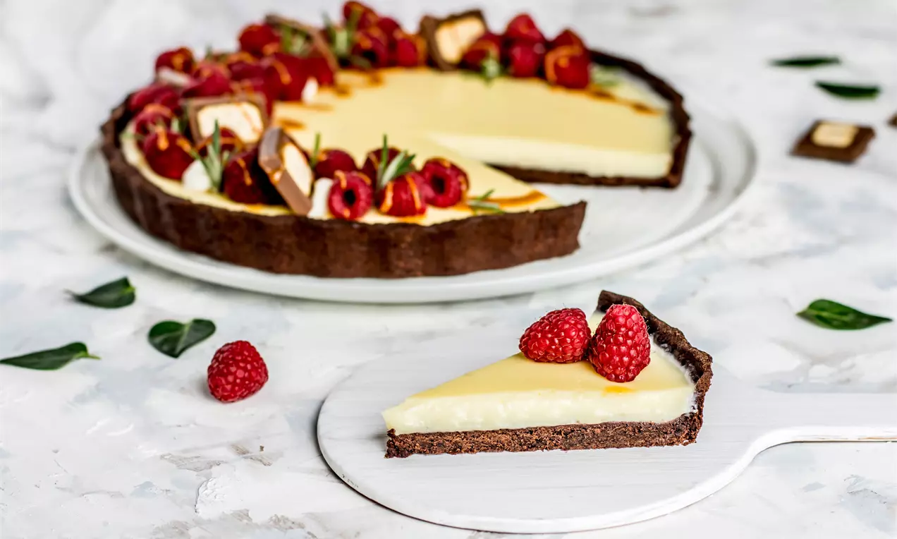 Шоколадов тарт с плътна кафява основа и крем Брюле с гладка светложълта текстура, украсен с цели малини и зеленки върху бяла чиния