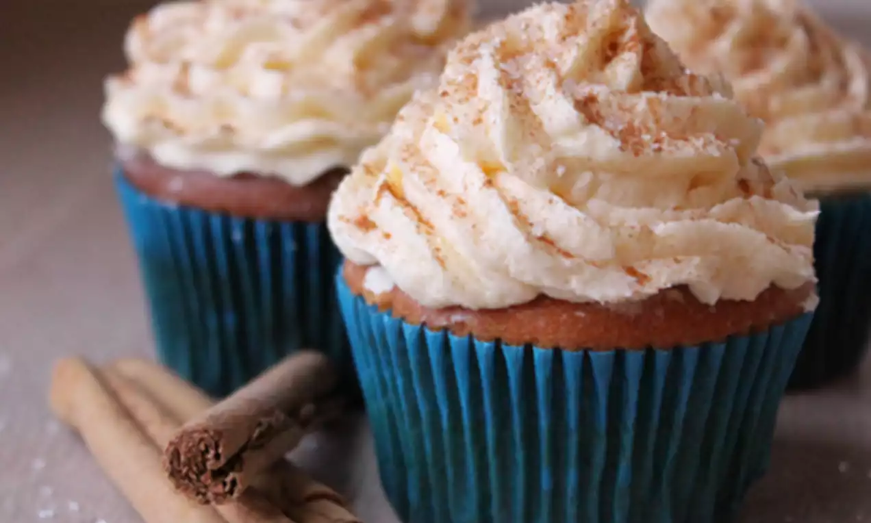 Kaneelkleurige cupcakes in blauwe papiertjes, bekroond met luchtige witte glazuur, bestoven met kaneel naast decoratieve kaneelstokjes