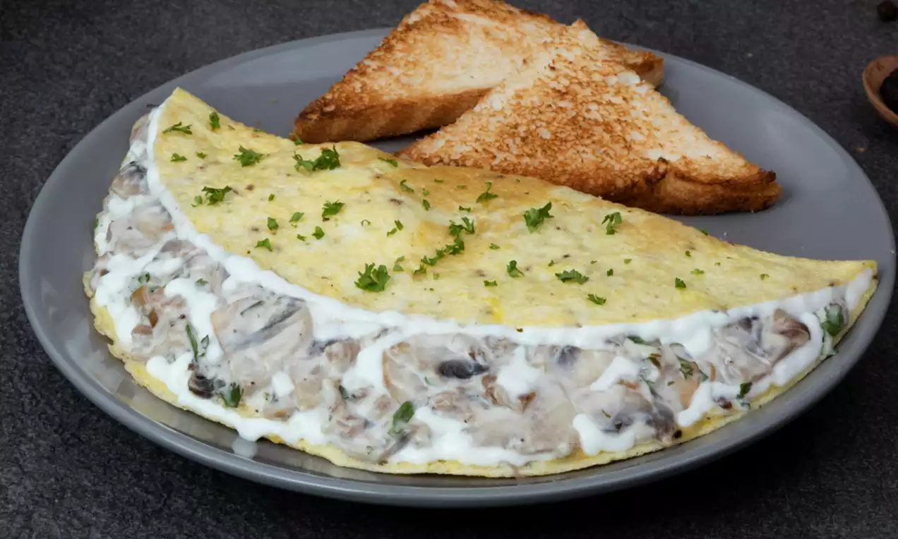 Golden-yellow folded omelette with a creamy mushroom filling, garnished with fresh parsley on a gray plate beside toasted bread slices