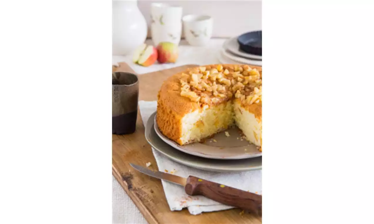 Ronde goudbruine cake met grove appelstukjes en kruimellaag bovenop, gepresenteerd op een grijs bord met een mes en een houten plank