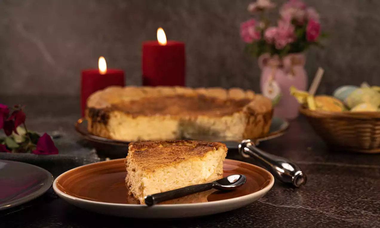 Pasca cu brânză rumenită și pufoasă, cu crusta aurie, feliată pe o farfurie maro, lângă lumânări roșii și decor de Paște pastelat