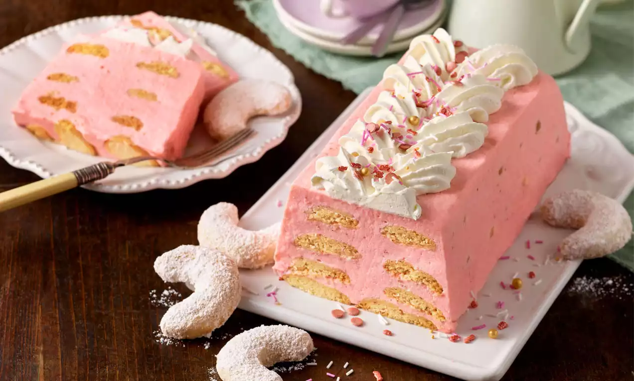 Rosafarbenes Erdbeerdessert mit eingearbeiteten Biskuitstücken als Rechteck auf weißer Platte, garniert mit Sahnetupfen, Streuseln und Vanillekipferln