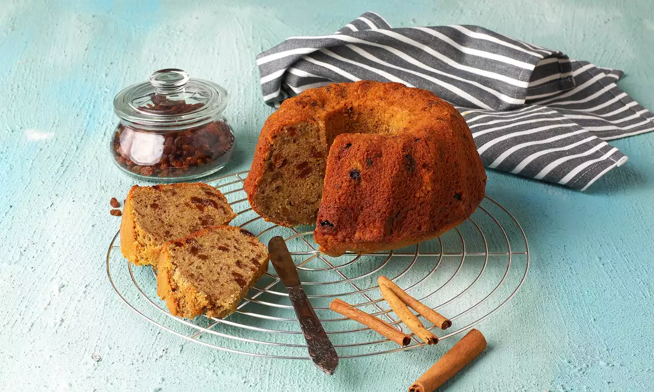 Altın kahverengi yüzeye sahip tarçınlı üzümlü cevizli kek, dilimlenmiş haliyle tel ızgara üzerinde, yanında tarçın çubukları ve cam kavanoz