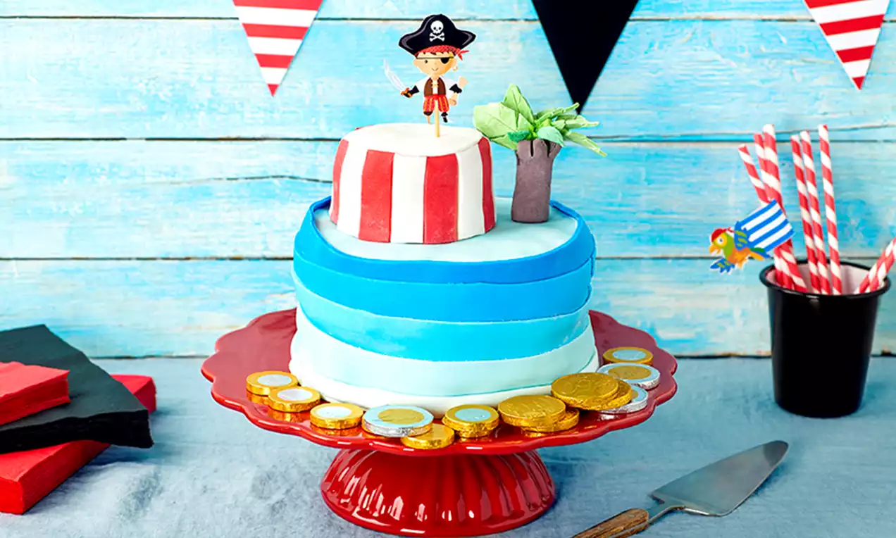 Tarta de dos pisos con franjas rojas y blancas, bandas azules degradadas, decorada con un pirata, palmera y monedas doradas sobre un plato rojo