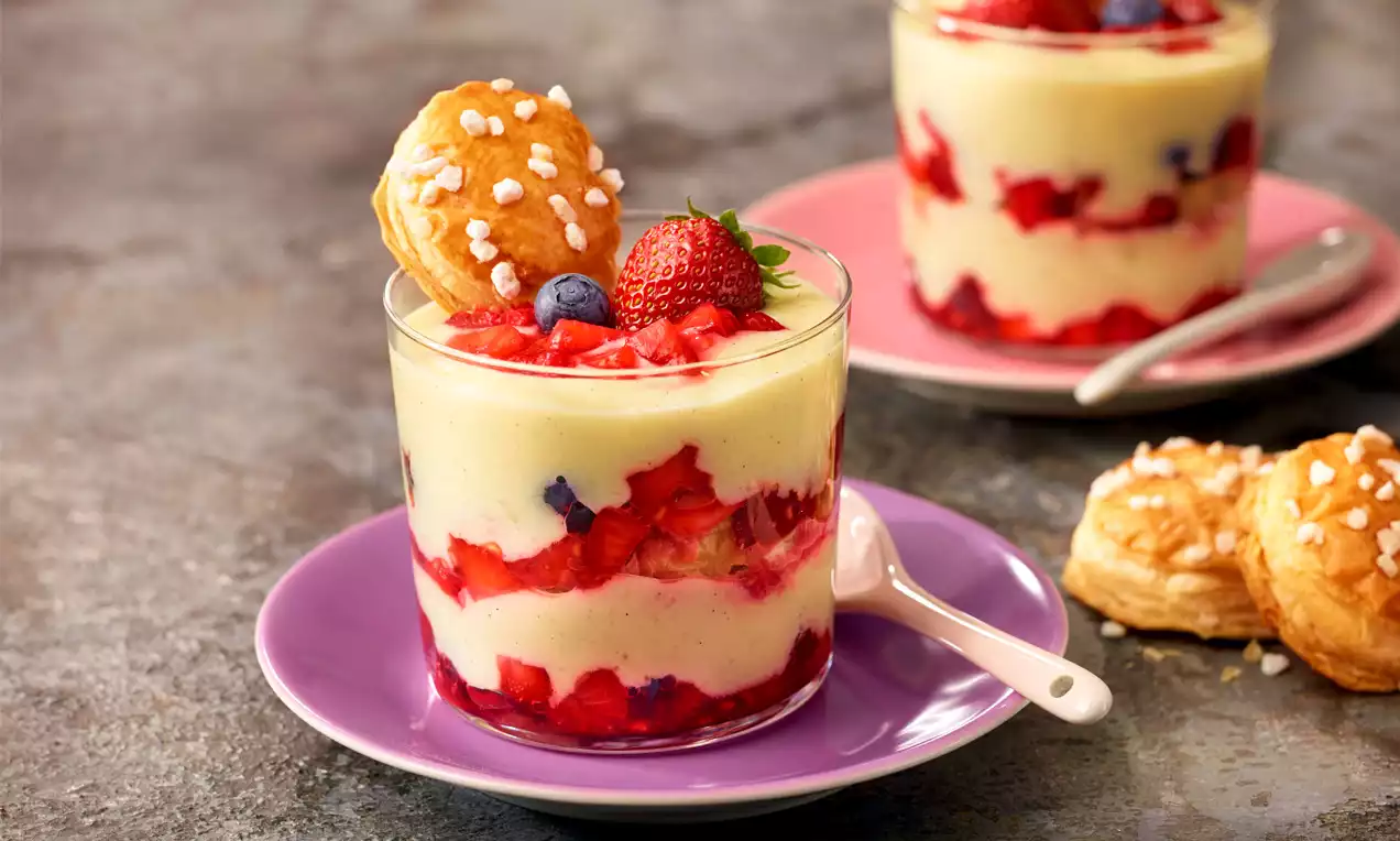 Schichtdessert mit gelber Creme, roten Beeren und einem gezuckerten Blätterteigstück in einem Glas auf einem lilafarbenen Teller