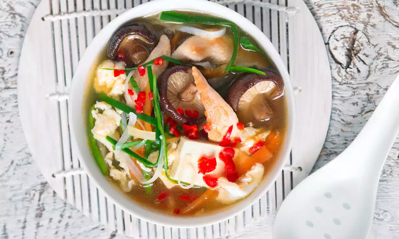 Světlý vývar s plátky masa, houbami, tofu, červenou chilli papričkou a zelenou cibulkou v bílé misce na dřevěném podkladu