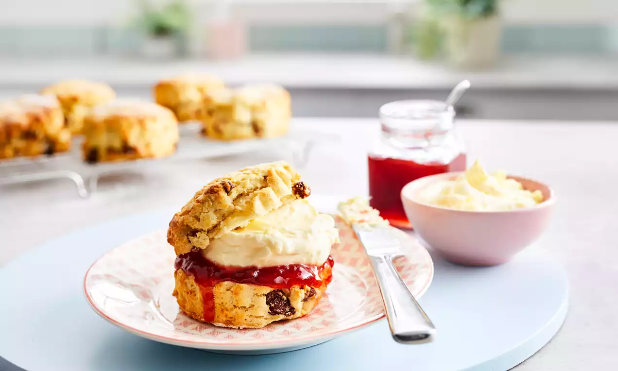 Golden gluten-free scone with a crumble texture, split and layered with glossy red jam and fluffy cream on a patterned plate